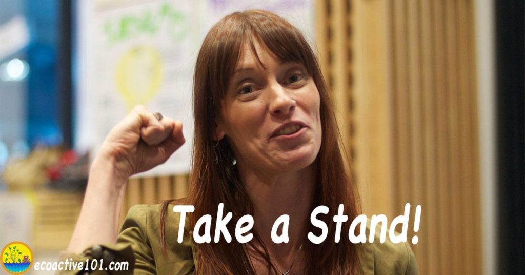 A young woman stands up in a meeting with raised fist, taking a stand for nuclear safety. The text says, “Take a stand!”